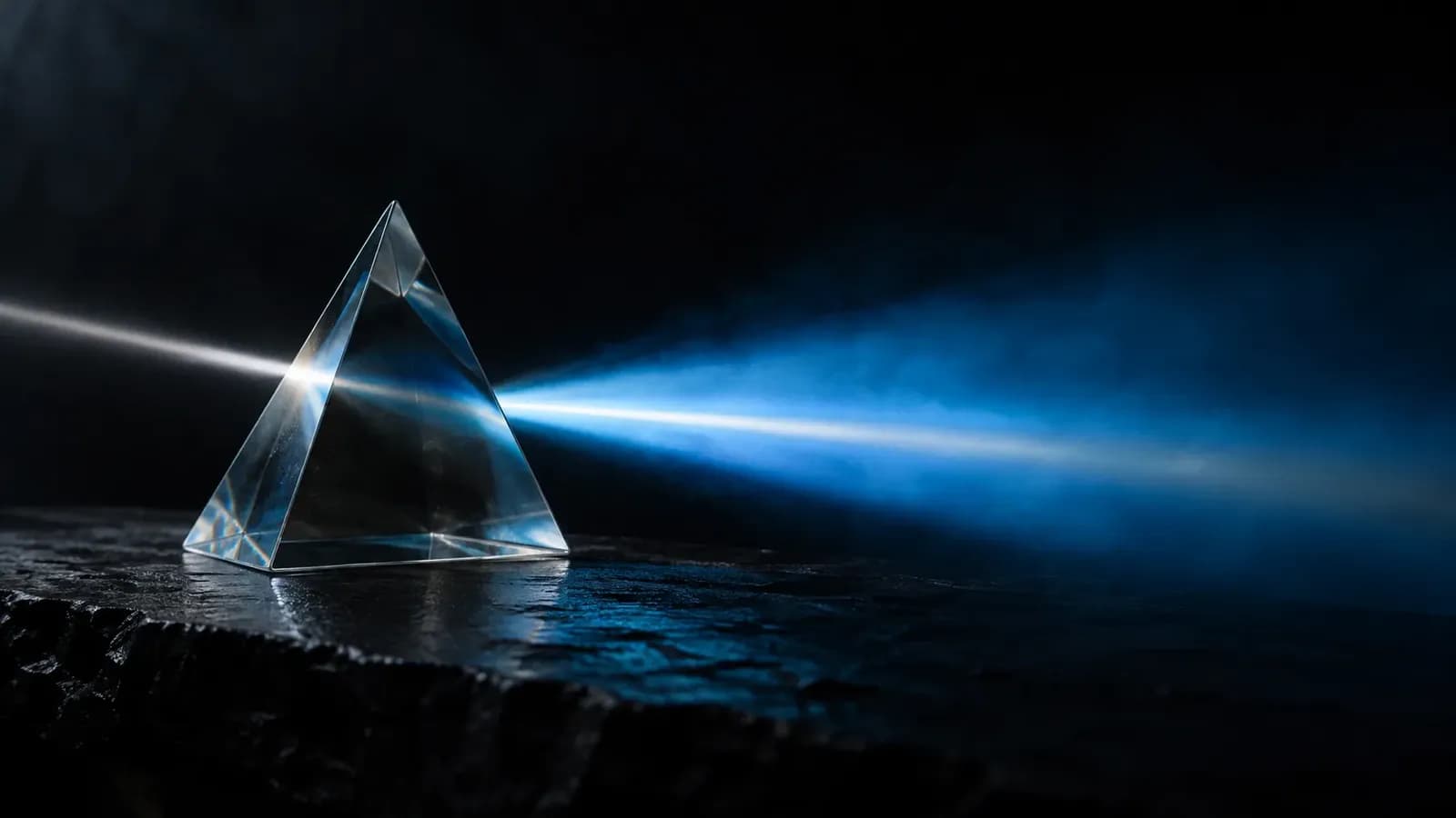 A clear glass prism on dark slate in a black room, where a thin white beam enters and spreads into a wide, soft sky-blue band of light with a brighter white core.
