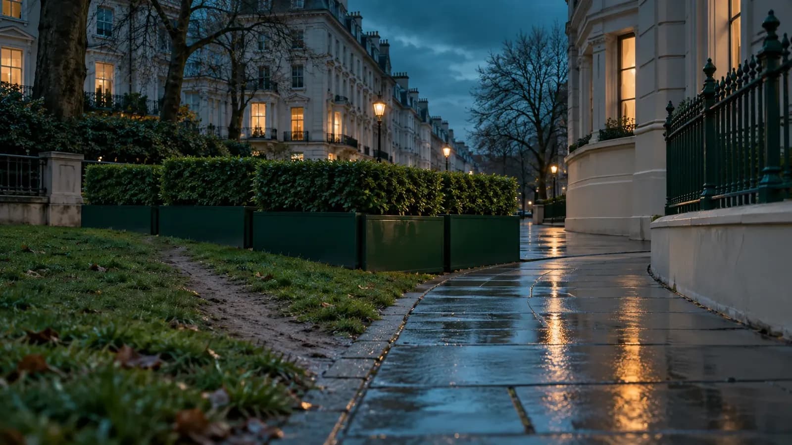 A blue-hour city street with wet pavement, where forest-green planters block a worn grass shortcut and a cream-stone footpath curves beside them, under a few warmly lit windows.