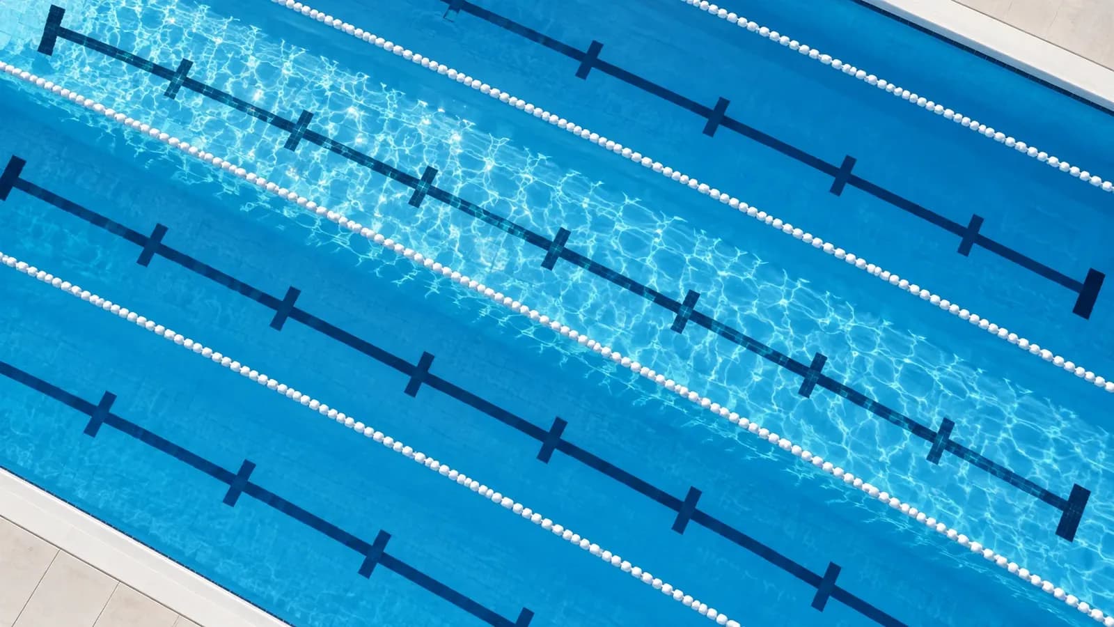 Overhead view of an empty swimming pool with sky blue water, white lane ropes and evenly spaced markers on the pool floor, lit by bright morning sun.