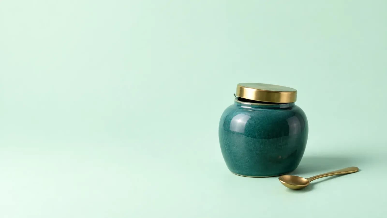 A deep teal ceramic jar with a brass lid turned slightly open, beside a small brass spoon, on a plain pale mint background with plenty of empty space.