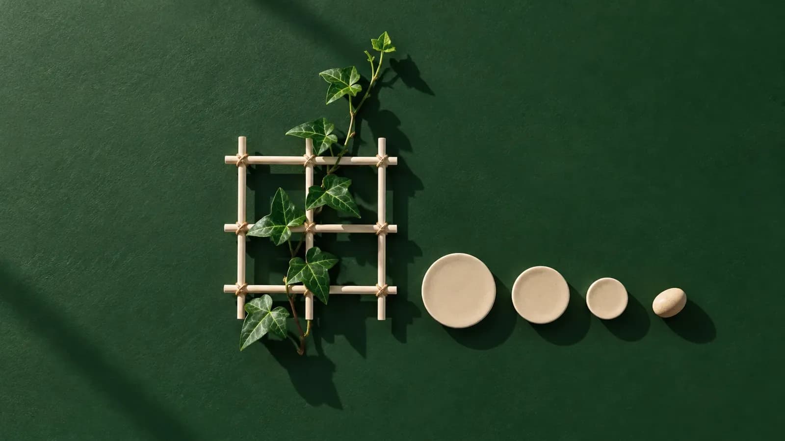 Top-down view of a small cream wooden trellis tied with twine on a forest green surface, with an ivy stem climbing through it beside a row of three shrinking cream discs and a pebble, under hard midday shadows.