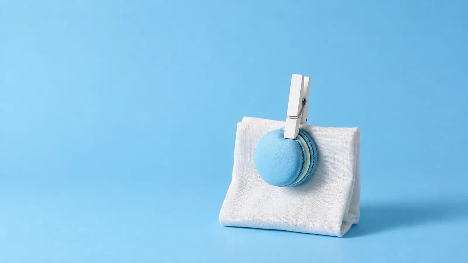A white clothes peg clips a pastel blue macaron to a folded white linen cloth on a plain sky blue backdrop, with plenty of empty space around them.